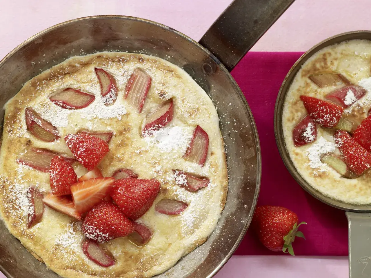 Zwei Pfannen mit frisch gebackenen Rhabarber-Pfannkuchen, garniert mit Erdbeeren und Puderzucker.