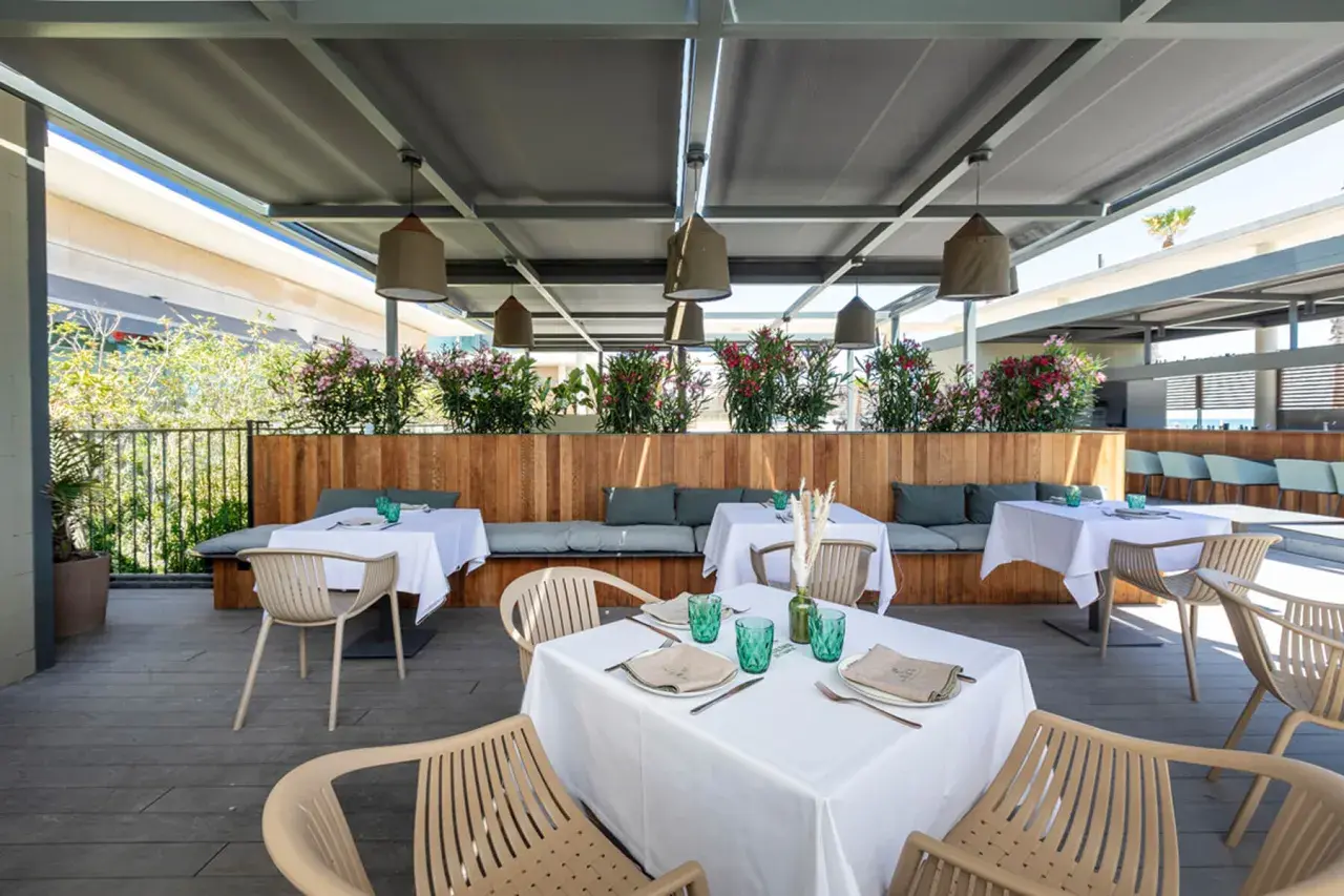 Terraza de la colonia de carmela con mesas puestas, asientos de madera y plantas floridas.