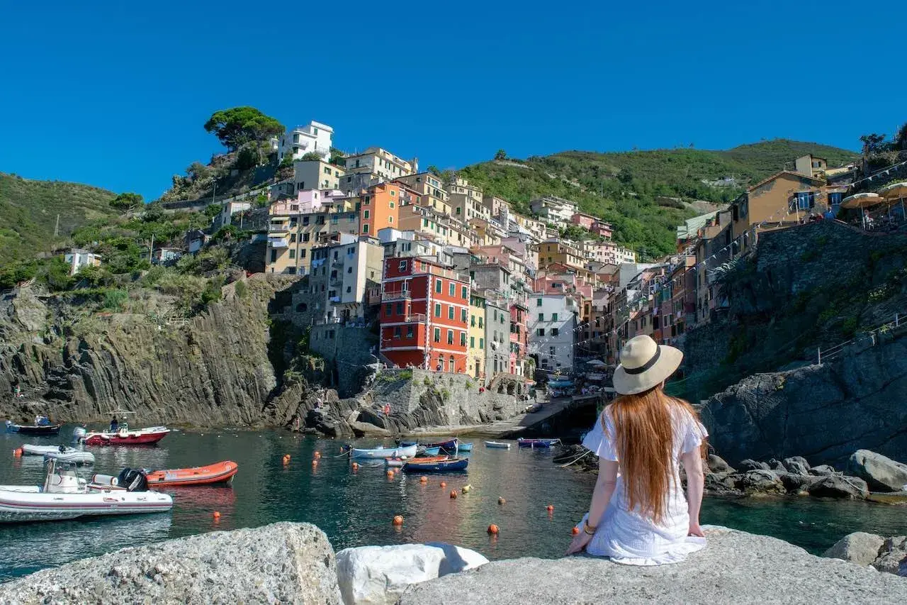 A woman in a white dress and hat gazes at the colorful houses of Riomaggiore, one of the best towns to stay in Cinque Terre, nestled on a cliffside overlooking the harbor.