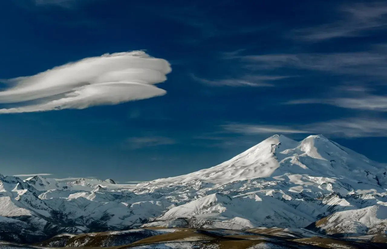 Pokryty śniegiem Elbrus, najwyższy szczyt Rosji, góruje nad ośnieżonymi pasmami górskimi pod błękitnym niebem z charakterystyczną chmurą.