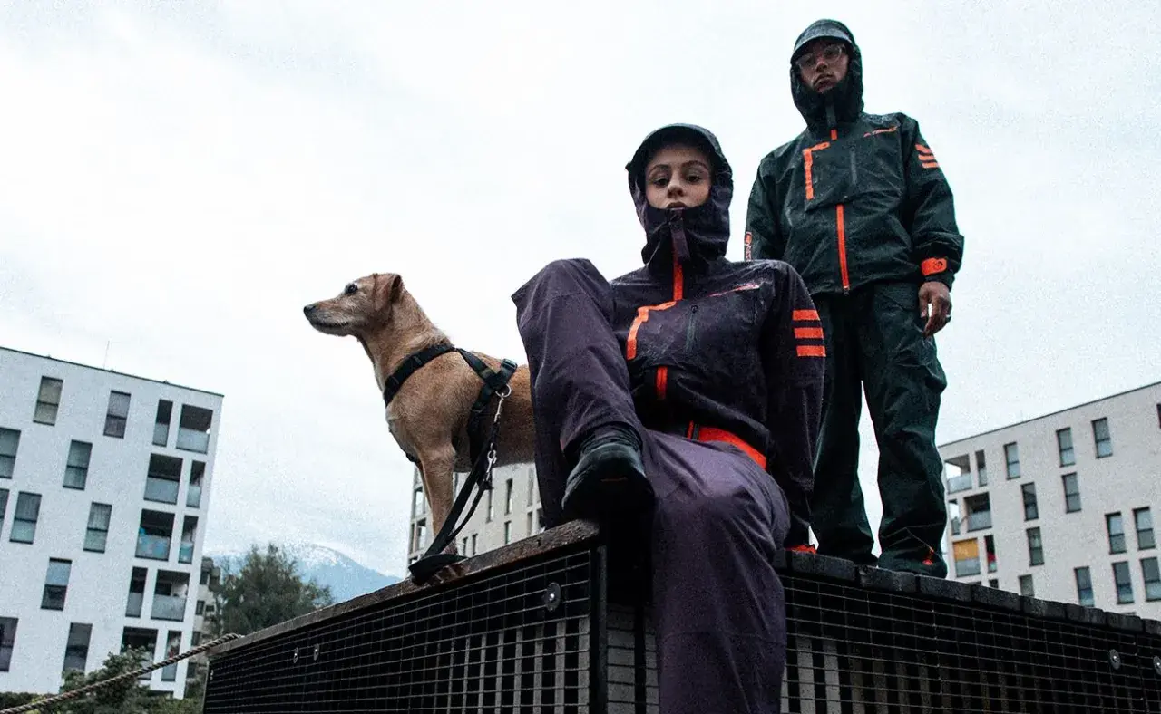 Zwei Personen in Gorpcore-Outfits und ein Hund stehen auf einem Metallgitter vor modernen Gebäuden.