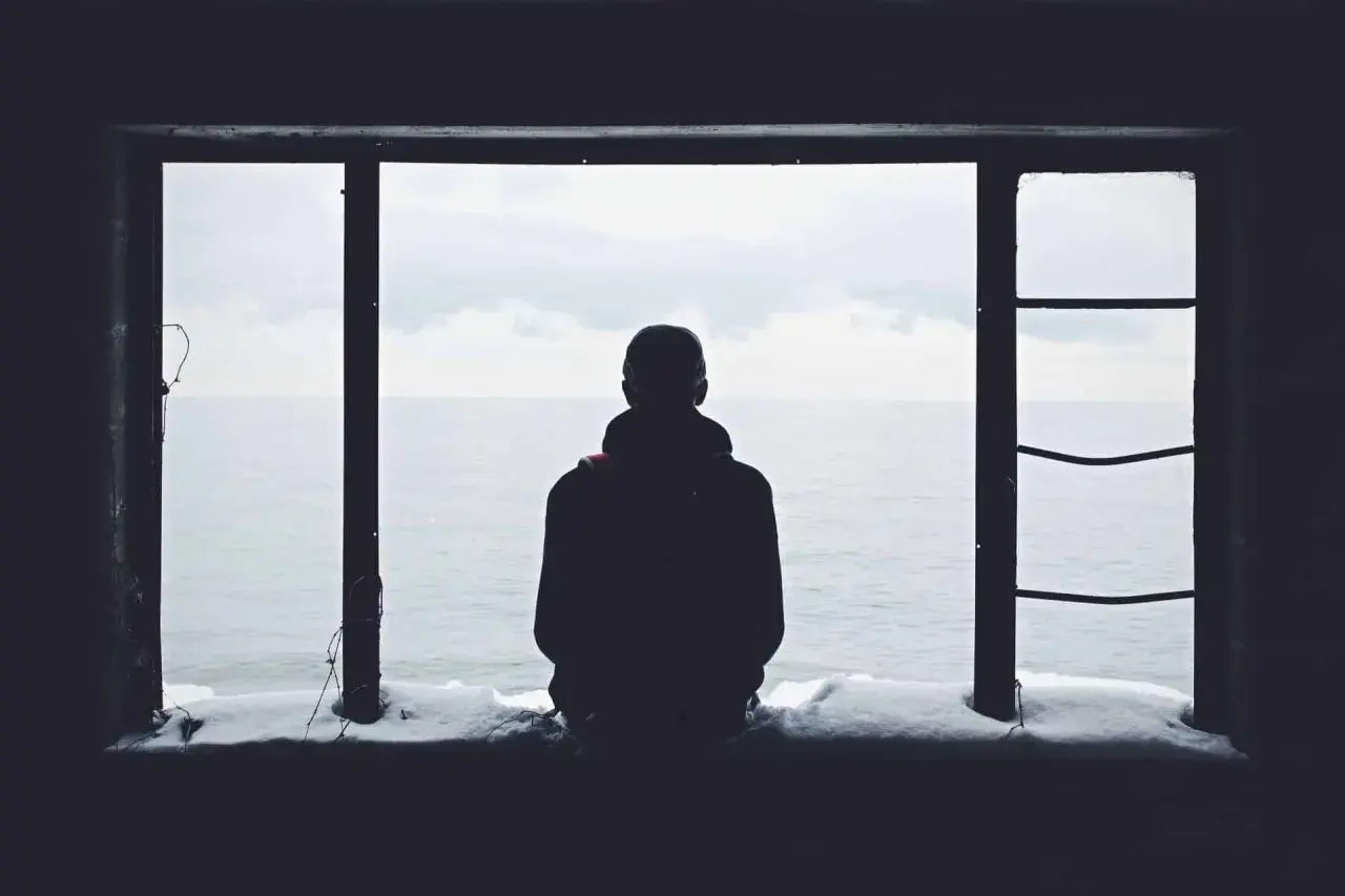 Eine Person blickt aus einem Fenster auf das Meer. Schnee liegt auf dem Sims. Das Bild vermittelt ein Gefühl von Einsamkeit.