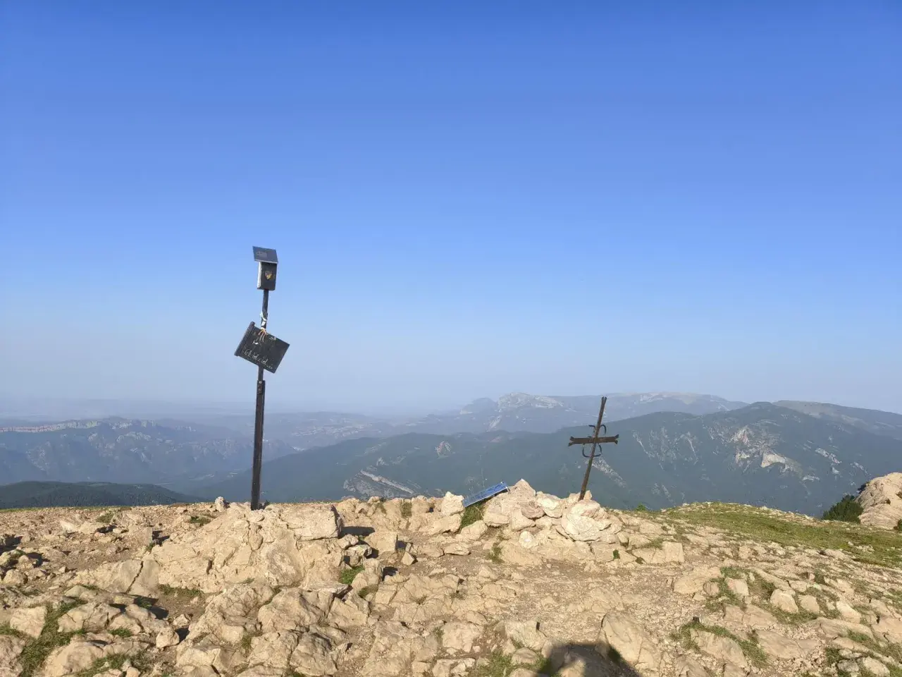 En el cap de la gallina pelada, un poste con placa y un sol solar vigilan el paisaje montañoso. A su lado, una cruz metálica se alza sobre rocas.