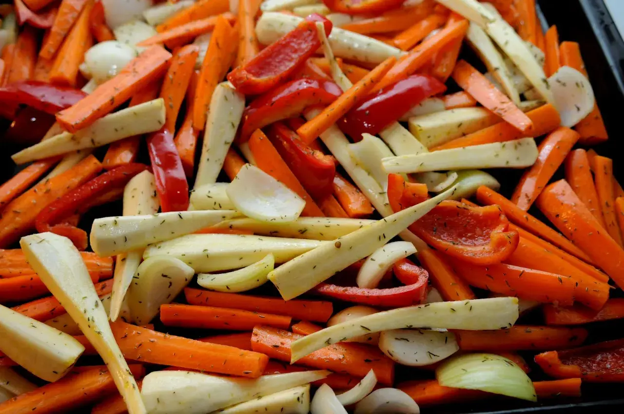 En ugnsplåt fylld med färgglada rotfrukter i ugn: morötter, palsternacka, paprika och lök, kryddade med örter.