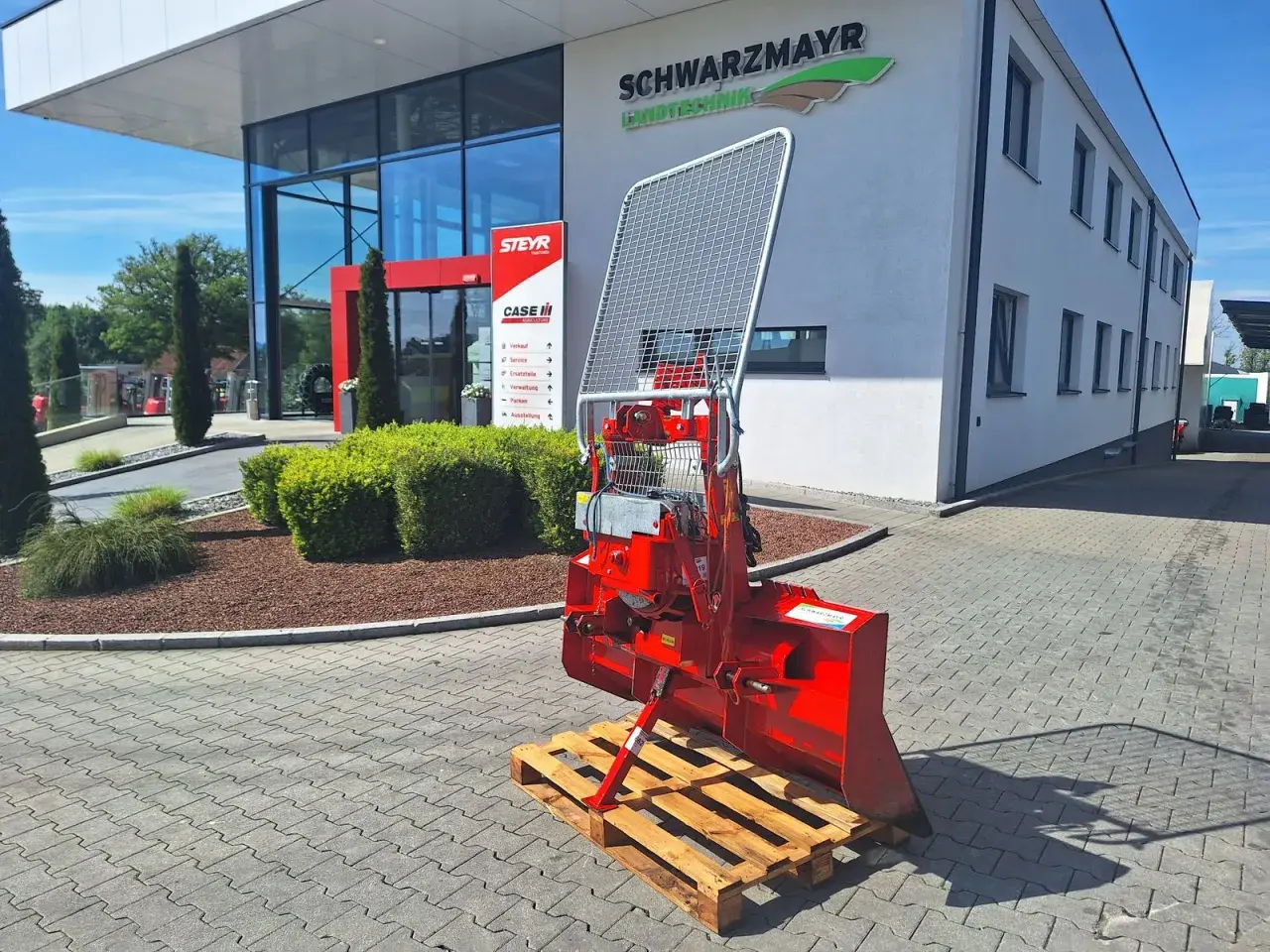 Roter Forstkran auf einer Palette vor einem Landtechnik-Gebäude. Ein robustes Gerät für den Einsatz im Wald, bereit für den Transport.