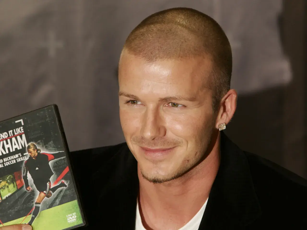 David Beckham, con su característico corte afeitado, sonríe mientras sostiene un DVD de sus habilidades de fútbol.