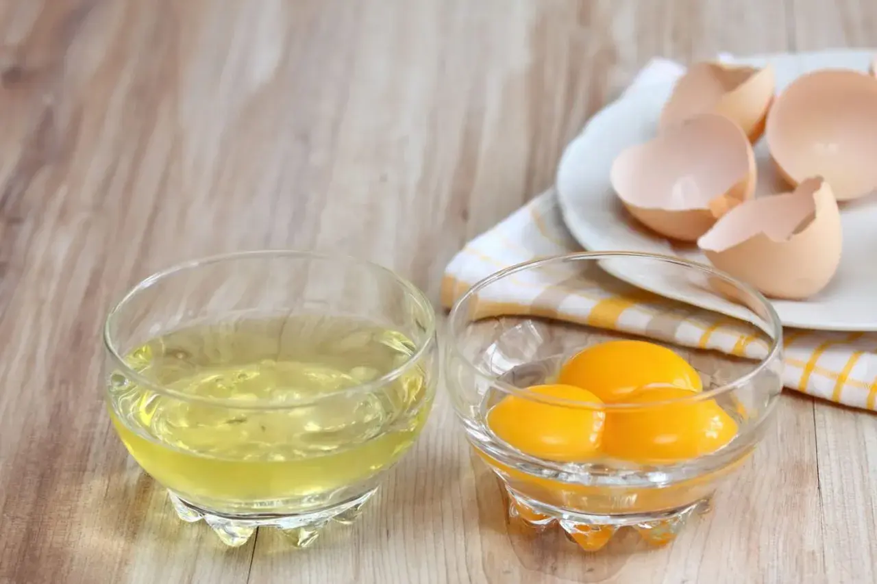 Deux bols en verre sur une table en bois : l'un contient du blanc d'œuf, l'autre des jaunes. Des coquilles d'œufs vides sont sur une assiette. Idéal pour la conservation du blanc d'œuf.