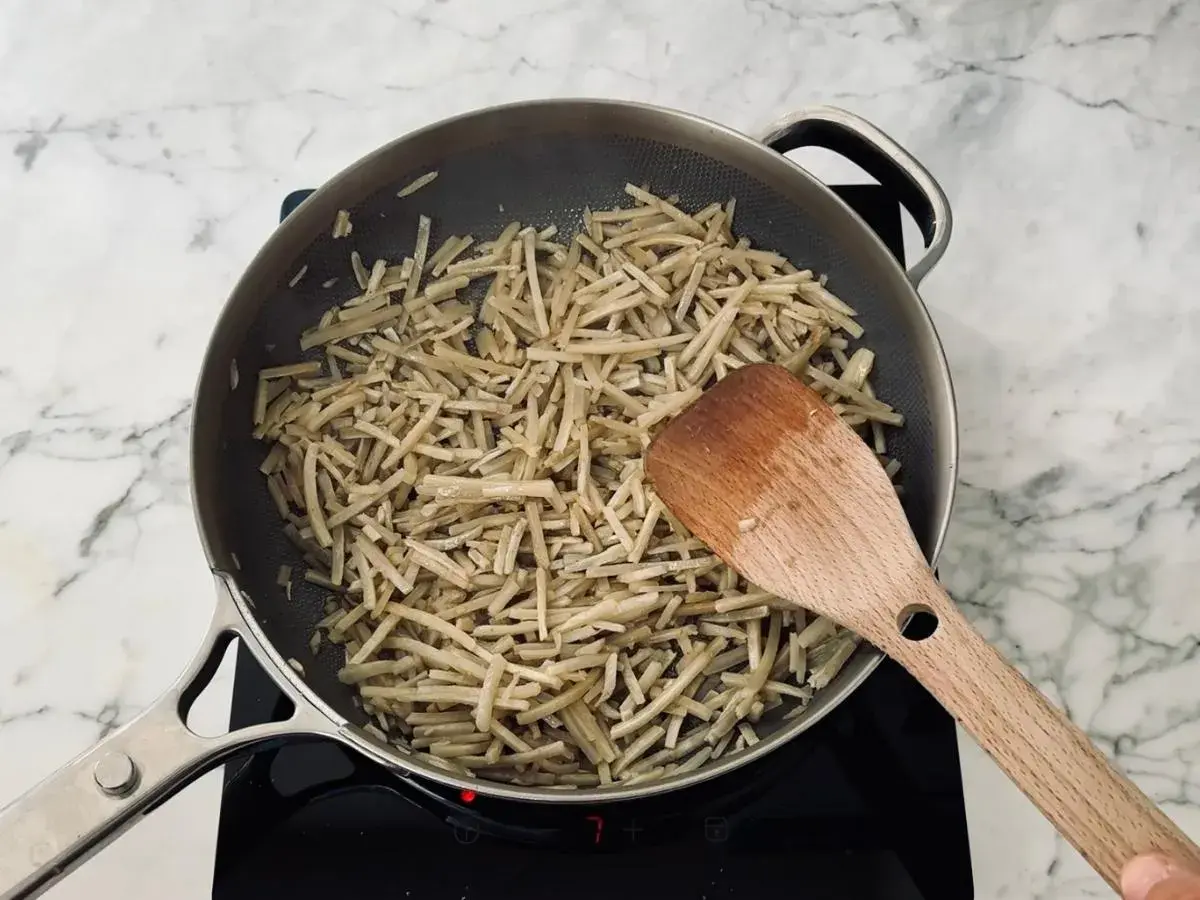 Strips of burdock root are being stir-fried in a pan, a key step in preparing a delicious kinpira recipe. A wooden spatula is used to toss the ingredients.