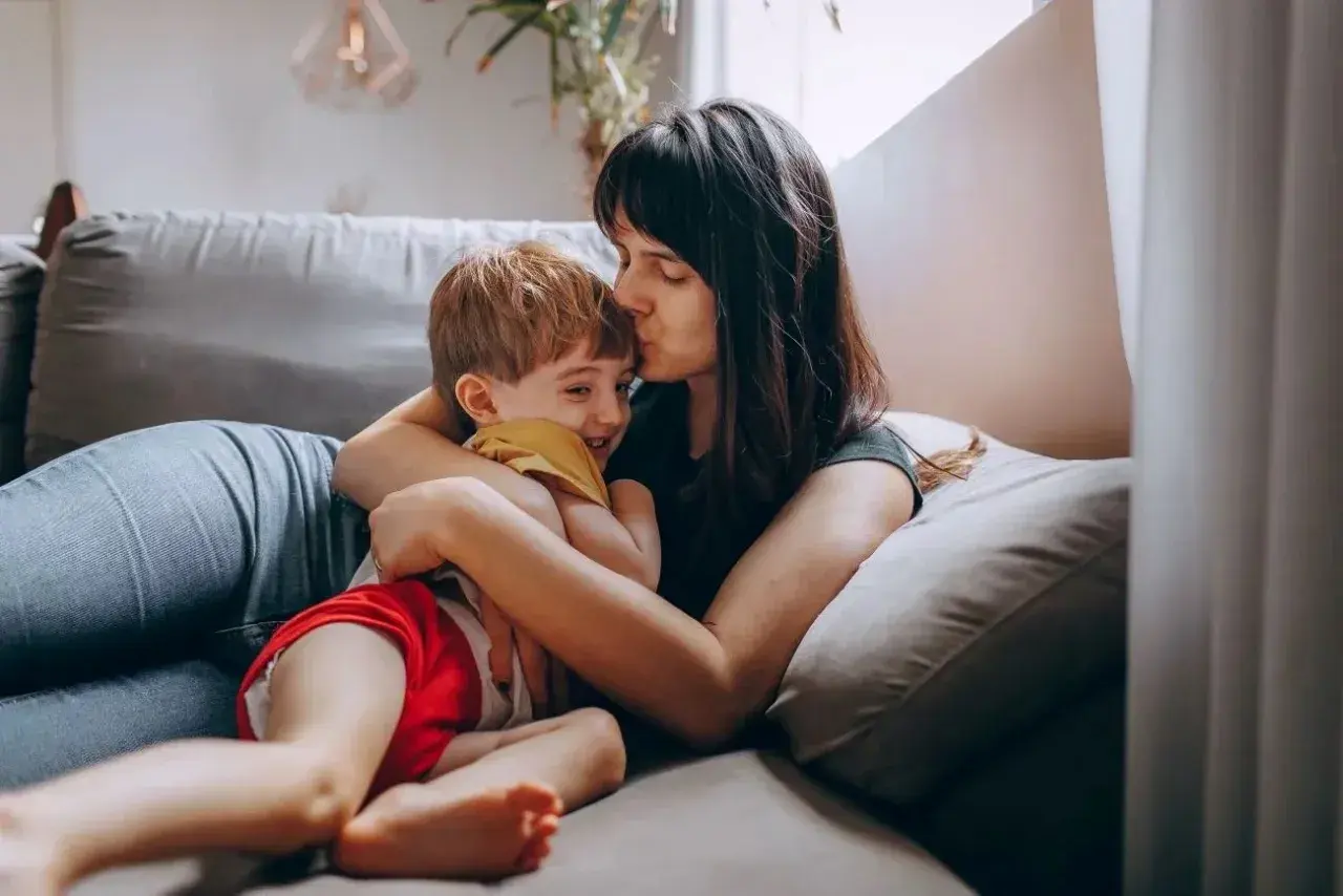 Mama całuje synka w czoło, przytulając go na kanapie. Dziecko śmieje się radośnie. Może dziś nie posyłam dziecka do przedszkola.