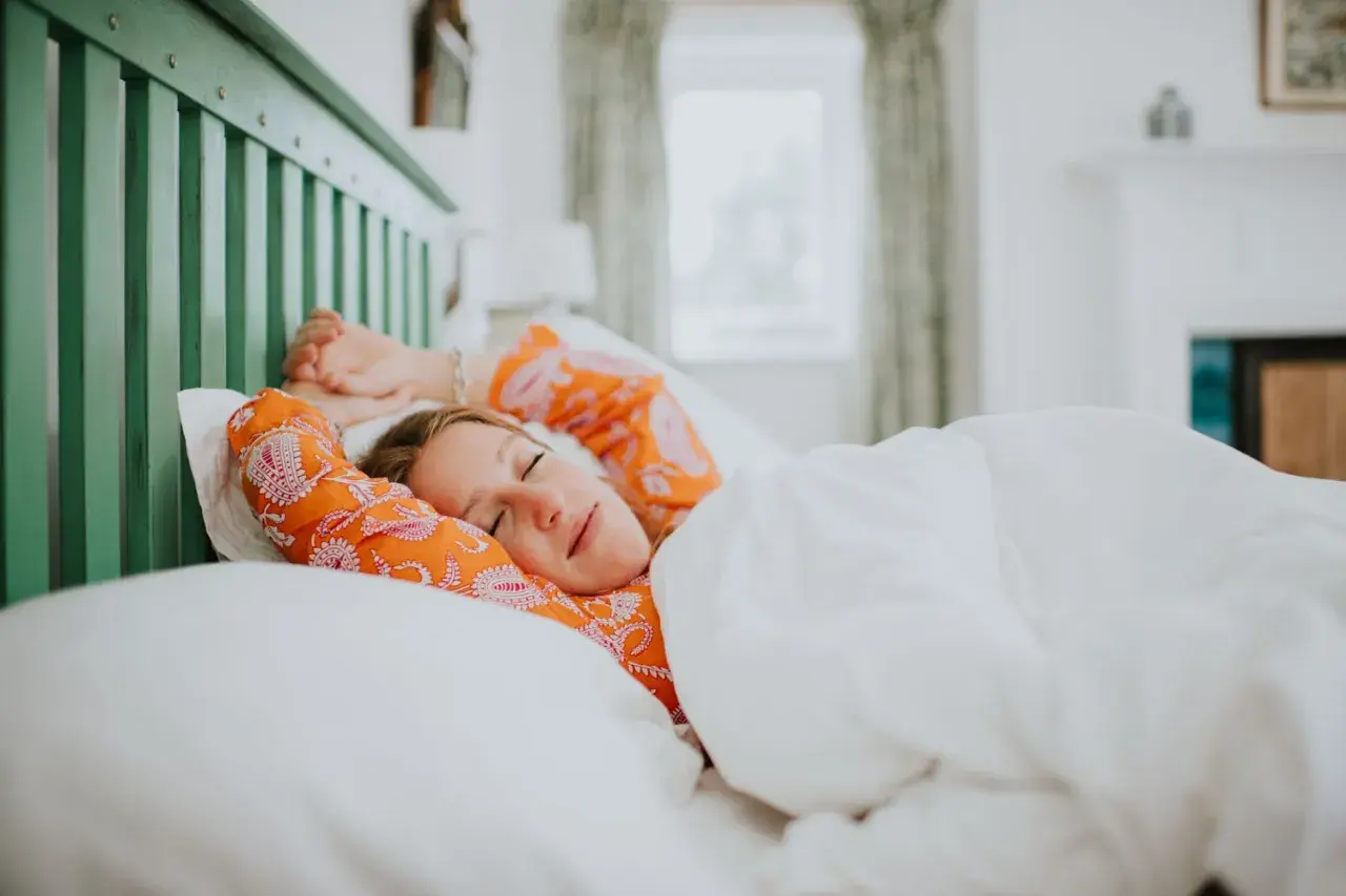 A woman in an orange patterned shirt stretches in bed, cozy under white sheets. She looks like she's getting just the right amount of light sleep.