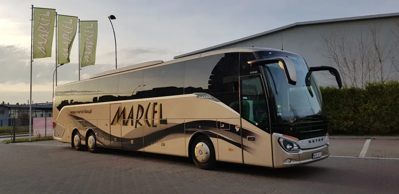 Autobus Marcel z napisem na boku, stojący na parkingu, z zielonymi flagami w tle.