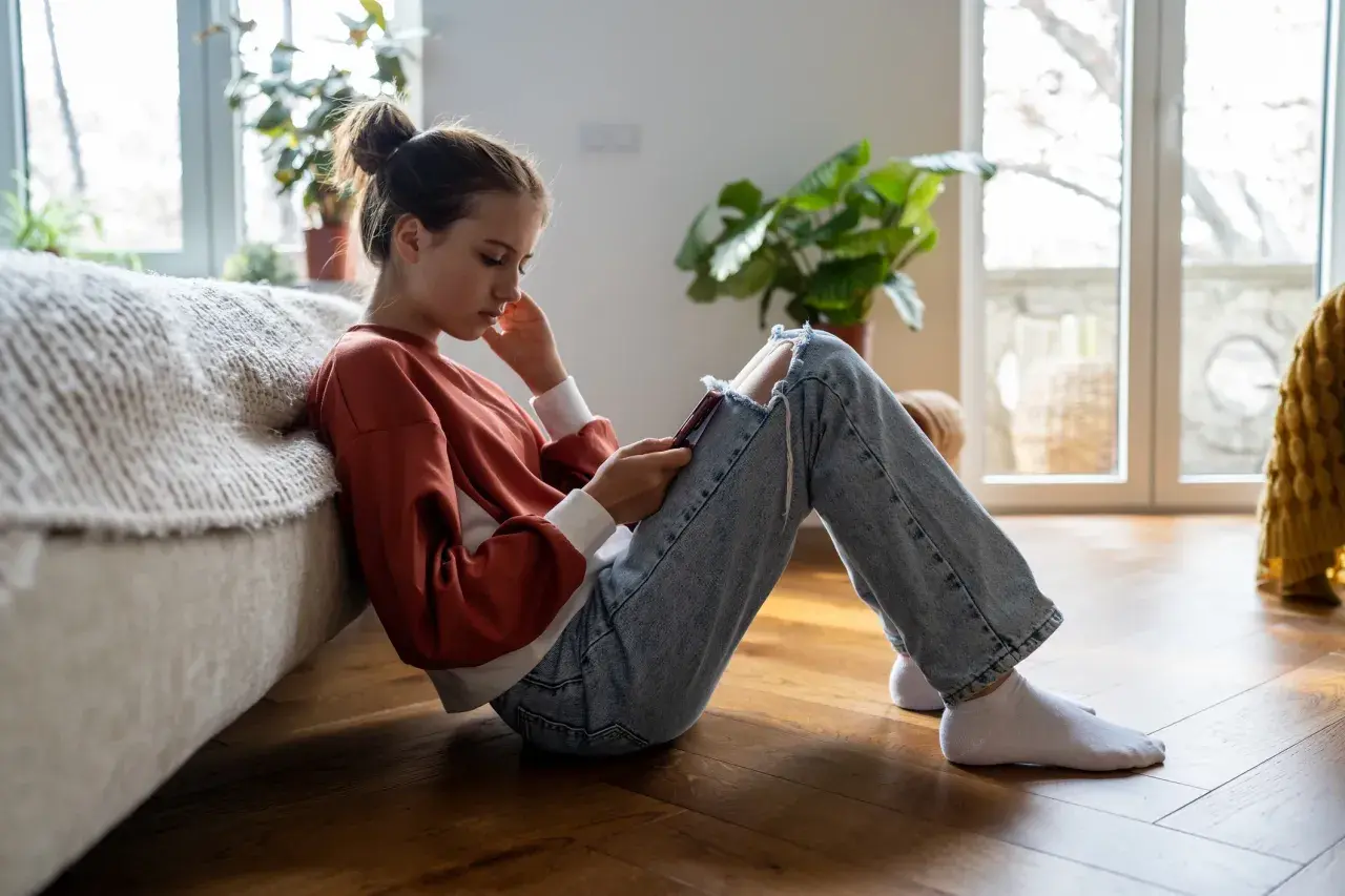 En flicka sitter på golvet, försjunken i sin telefon. Hennes hållning och ansiktsuttryck kan antyda depression hos barn.