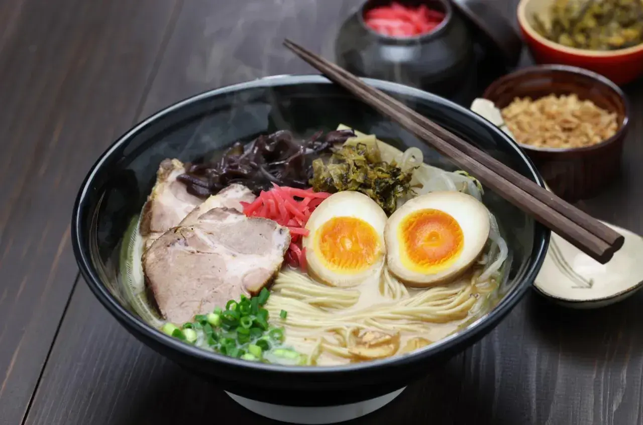 Pyszny ramen co to? Miska parującego bulionu z makaronem, plastrami wieprzowiny, jajkiem na pół, szczypiorkiem i marynowanymi dodatkami.