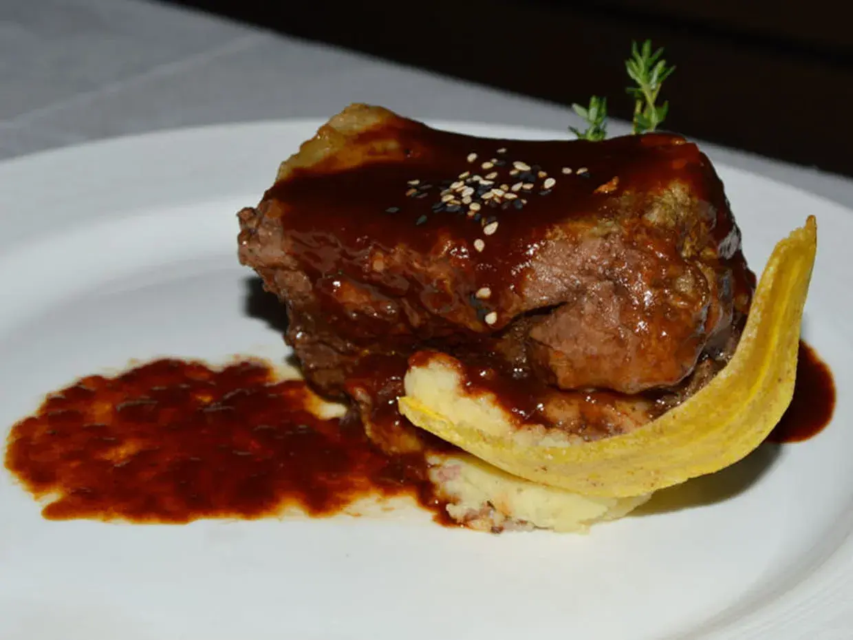 Un plat de ribs de boeuf barbecue, nappé d'une sauce foncée, servi avec une purée et une tranche de banane plantain frite.
