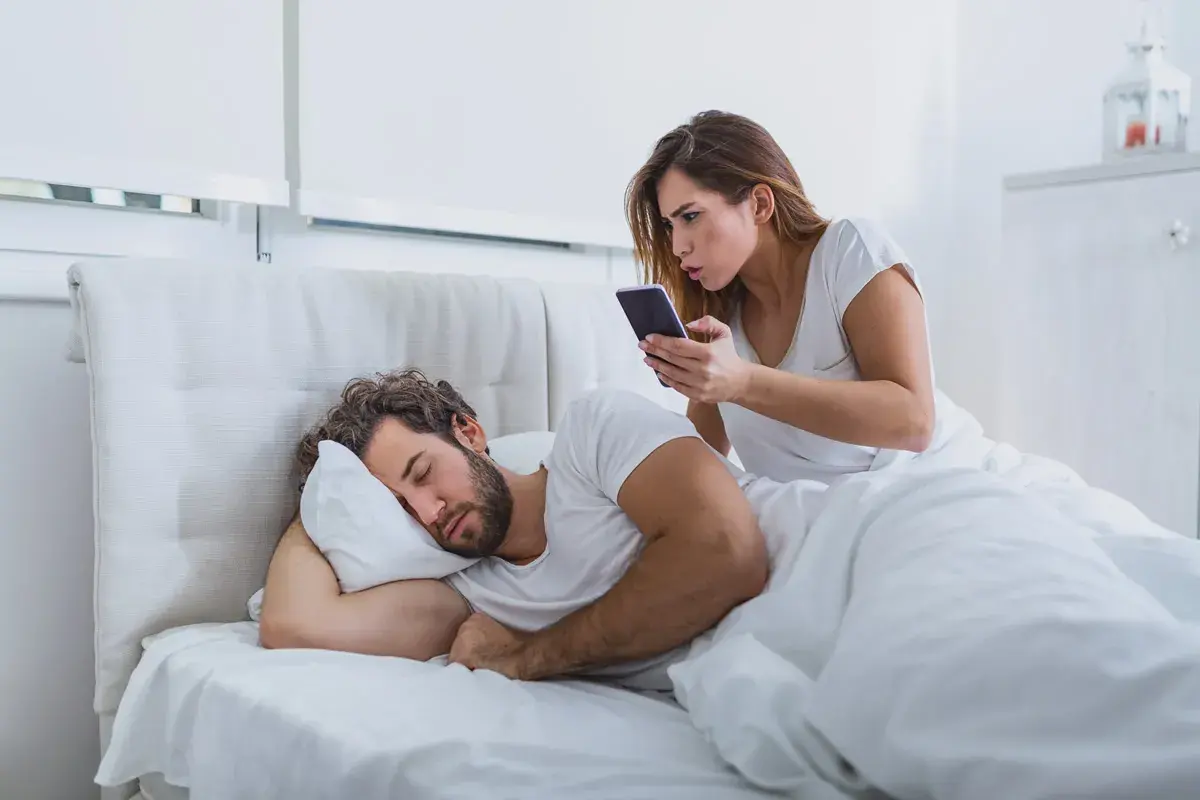 Donna preoccupata controlla il telefono del partner addormentato, un classico esempio di psicologia della sfiducia.