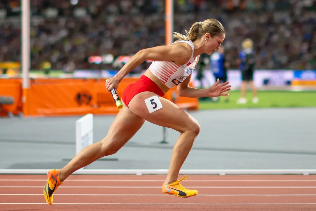 Zawodniczka biegnie z pałeczką w sztafecie 4x100 kobiet. Numer 5 na udzie.