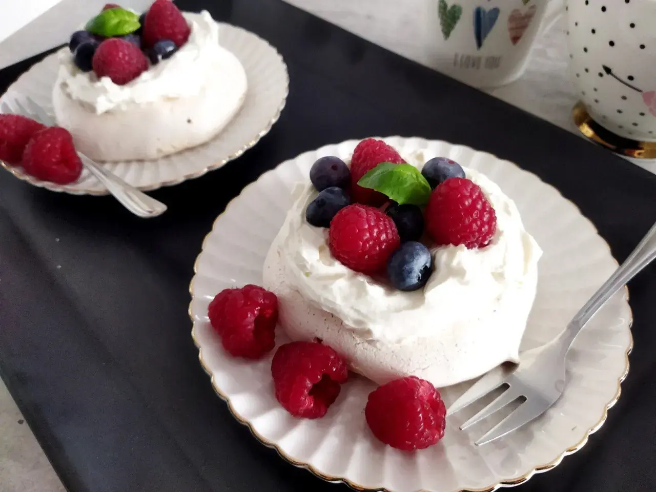 Dwa torty bezowe pavlova z bitą śmietaną, malinami i borówkami na talerzykach.
