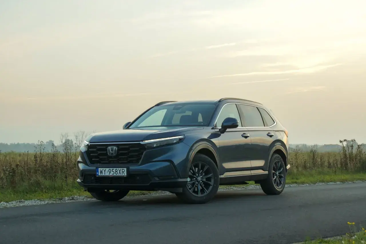 Ciemnoniebieski Honda CR-V Hybrid na tle zachodzącego słońca, zaparkowany na poboczu drogi.