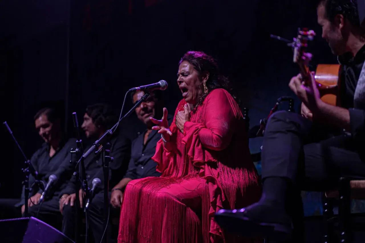 Una cantante colombiana flamenco, con un vestido rojo vibrante, canta apasionadamente acompañada por guitarristas.