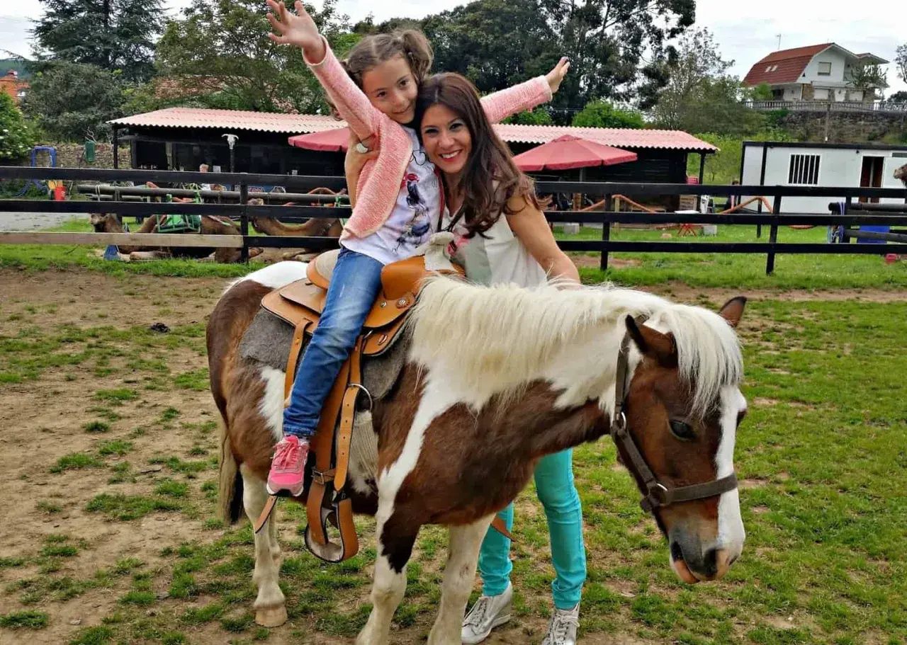 Niña feliz en un poni, disfrutando de una ruta a caballo en Cantabria. Madre sonriente la acompaña.
