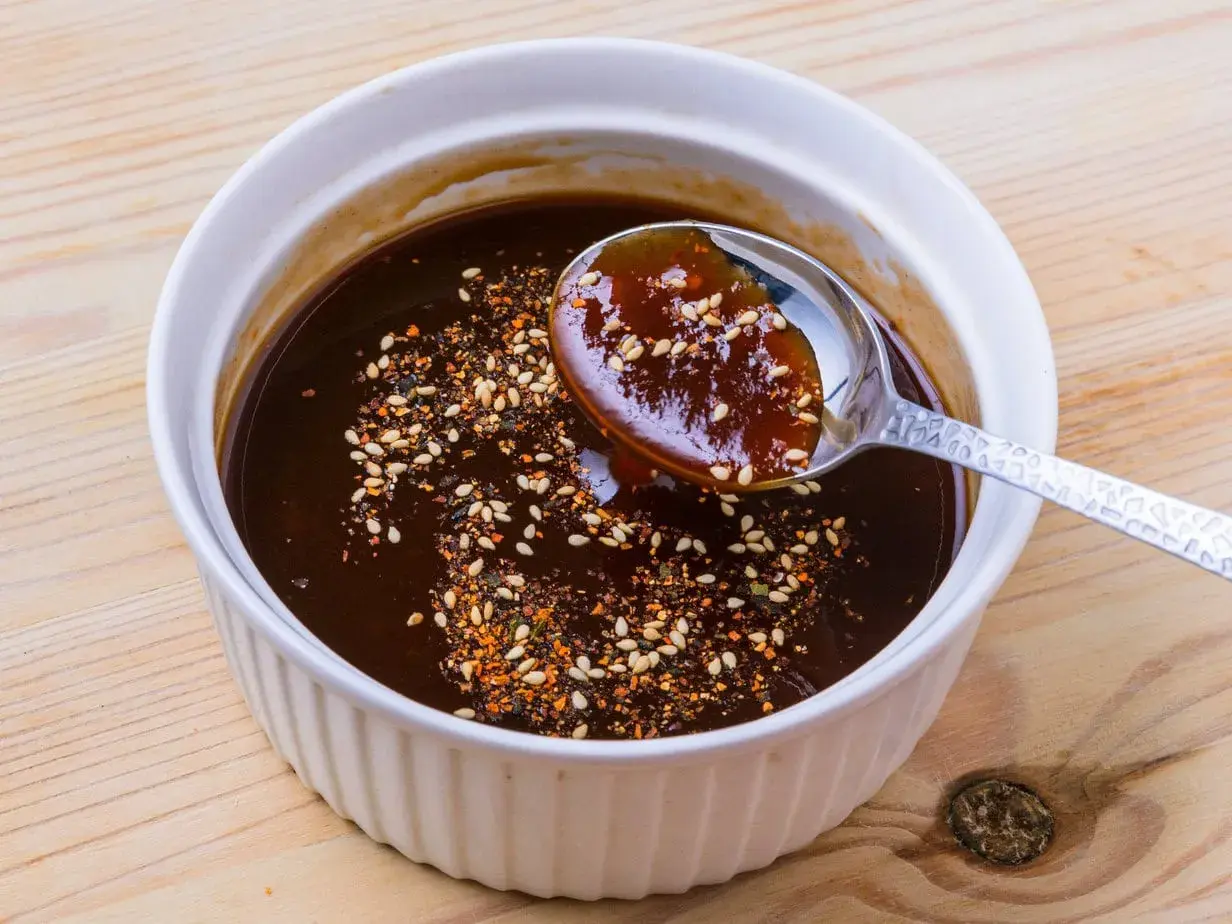 A spoon scoops rich, dark okonomiyaki sauce from a white bowl, sprinkled with sesame seeds and chili flakes. This is how to make okonomiyaki sauce.