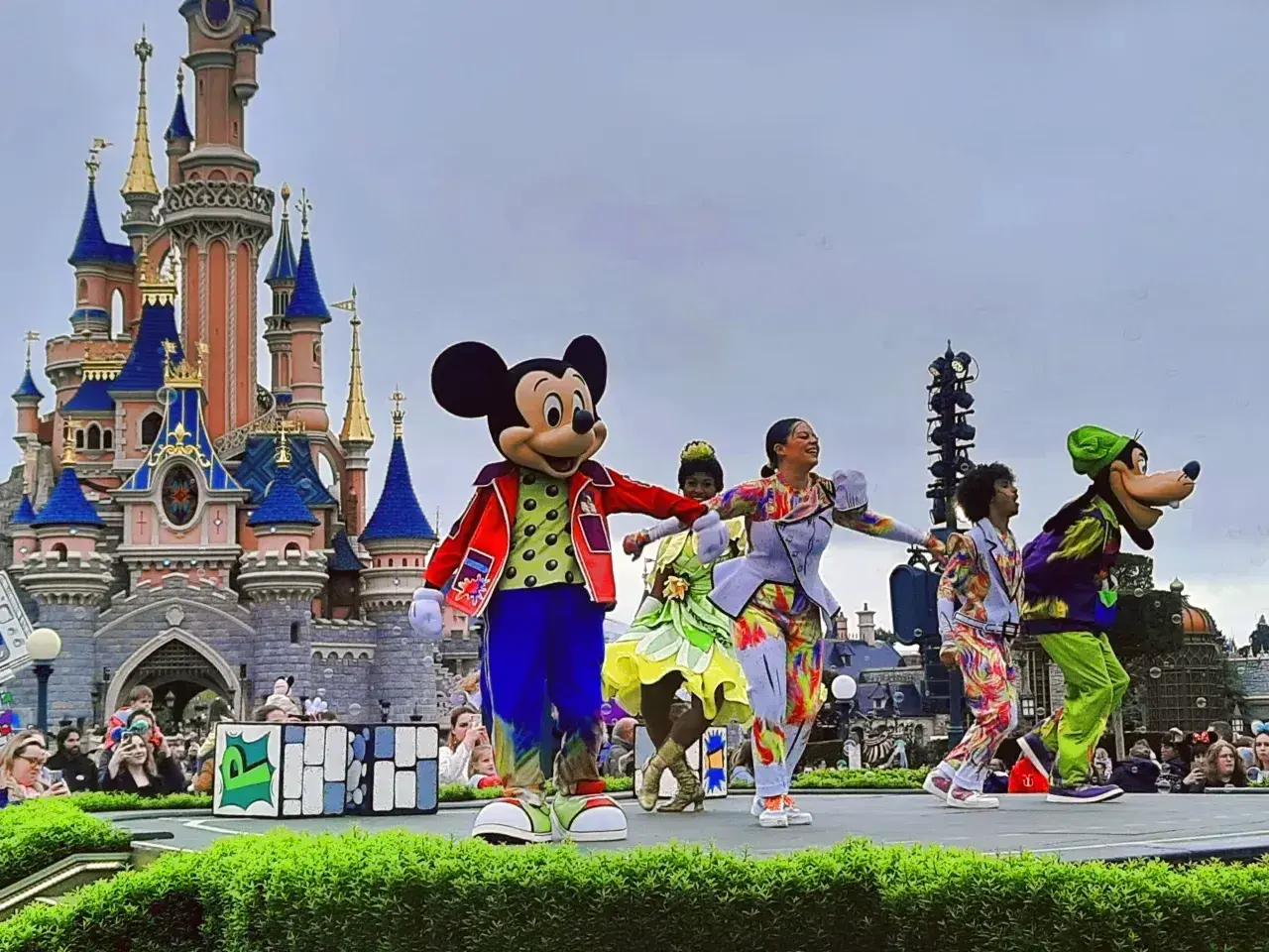 Myszka Miki i Goofy na scenie przed zamkiem w Disneyland Paris, otoczeni tancerzami w kolorowych strojach. Niezapomniane atrakcje Disneyland Paris!