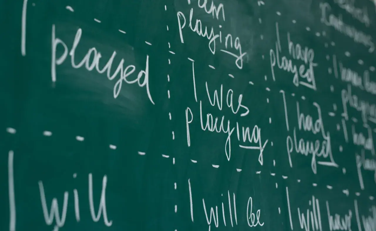 Scrittura a mano su lavagna verde: "I played", "I was playing", "I have played", "I will play". Esercizi di grammatica inglese, con focus sui tempi verbali.