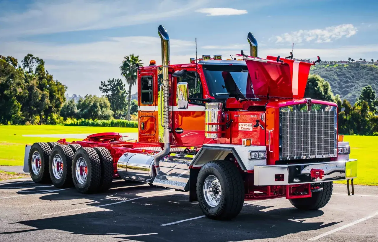 Un impressionnant Kenworth, un camion américain rouge vif, brille sous le soleil. Ses chromes étincelants et ses échappements imposants annoncent sa puissance.