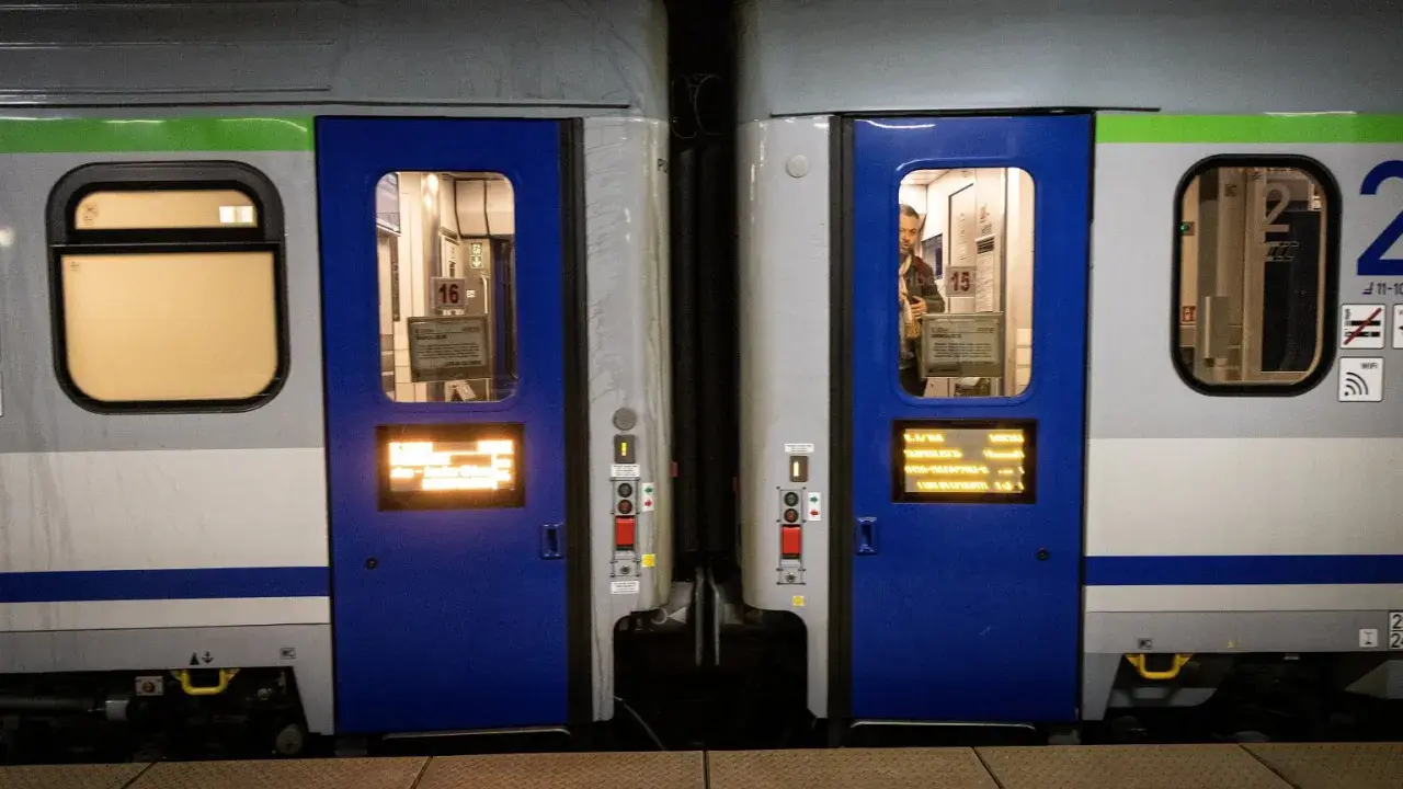 Dwa wagony kolejowe z niebieskimi drzwiami i oknami. Widać numery wagonów 16 i 15. Różne rodzaje wagonów czekają na pasażerów.