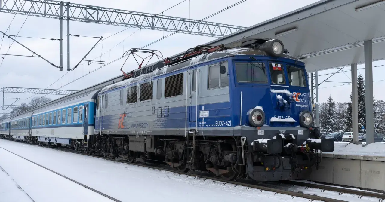 Lokomotywa EU07-421 PKP Intercity z pociągiem na tle zimowego krajobrazu. Widać elementy sieci trakcyjnej i peron.