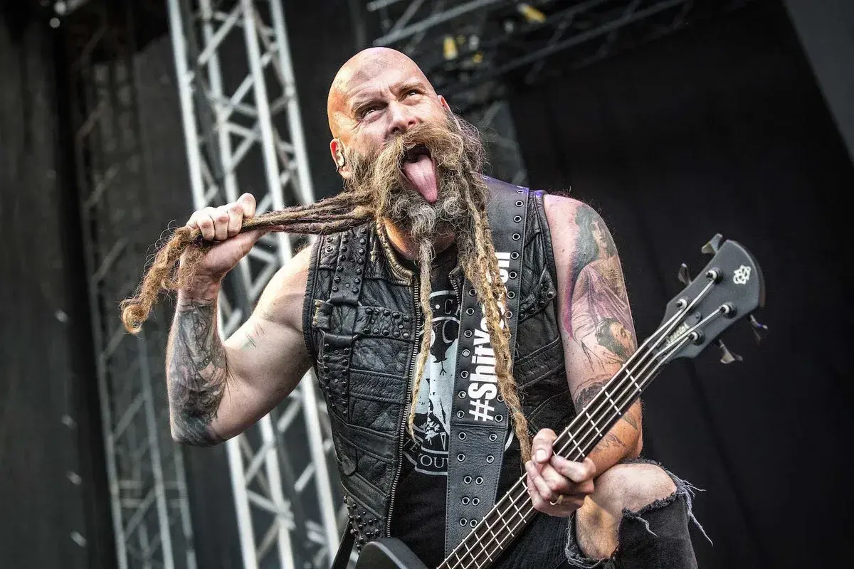 Chris Kael, basista z długą, zaplecioną brodą, wyciąga język podczas koncertu, trzymając gitarę basową.