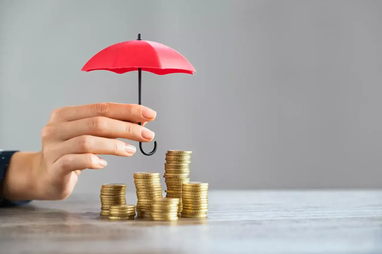 Une main protège des piles de pièces avec un petit parapluie rouge, symbolisant l'assurance PTZ.