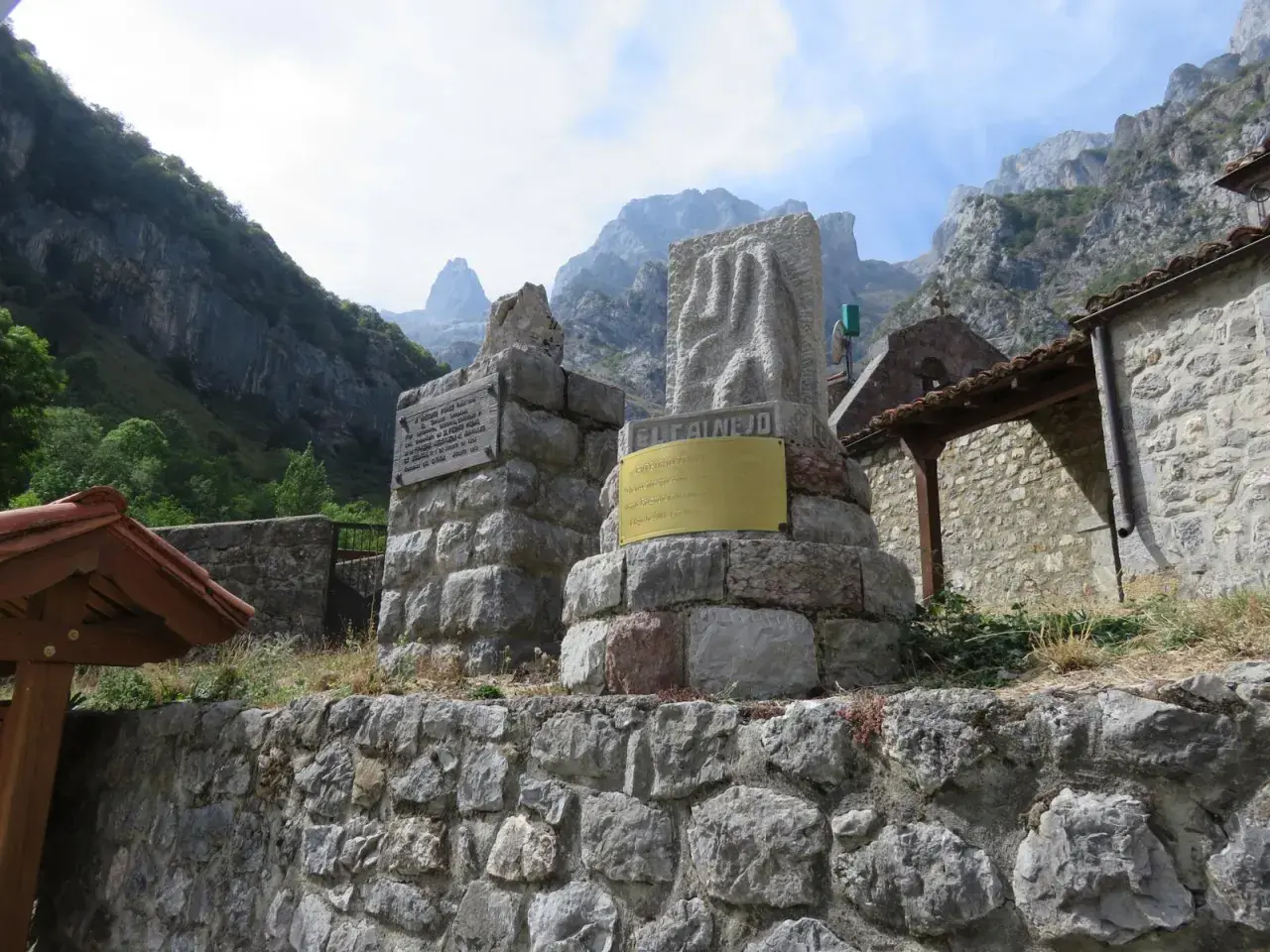 Monumento en piedra con inscripciones y un relieve, con montañas al fondo. El cartel dice "EL CAIN DE ARRIBA".