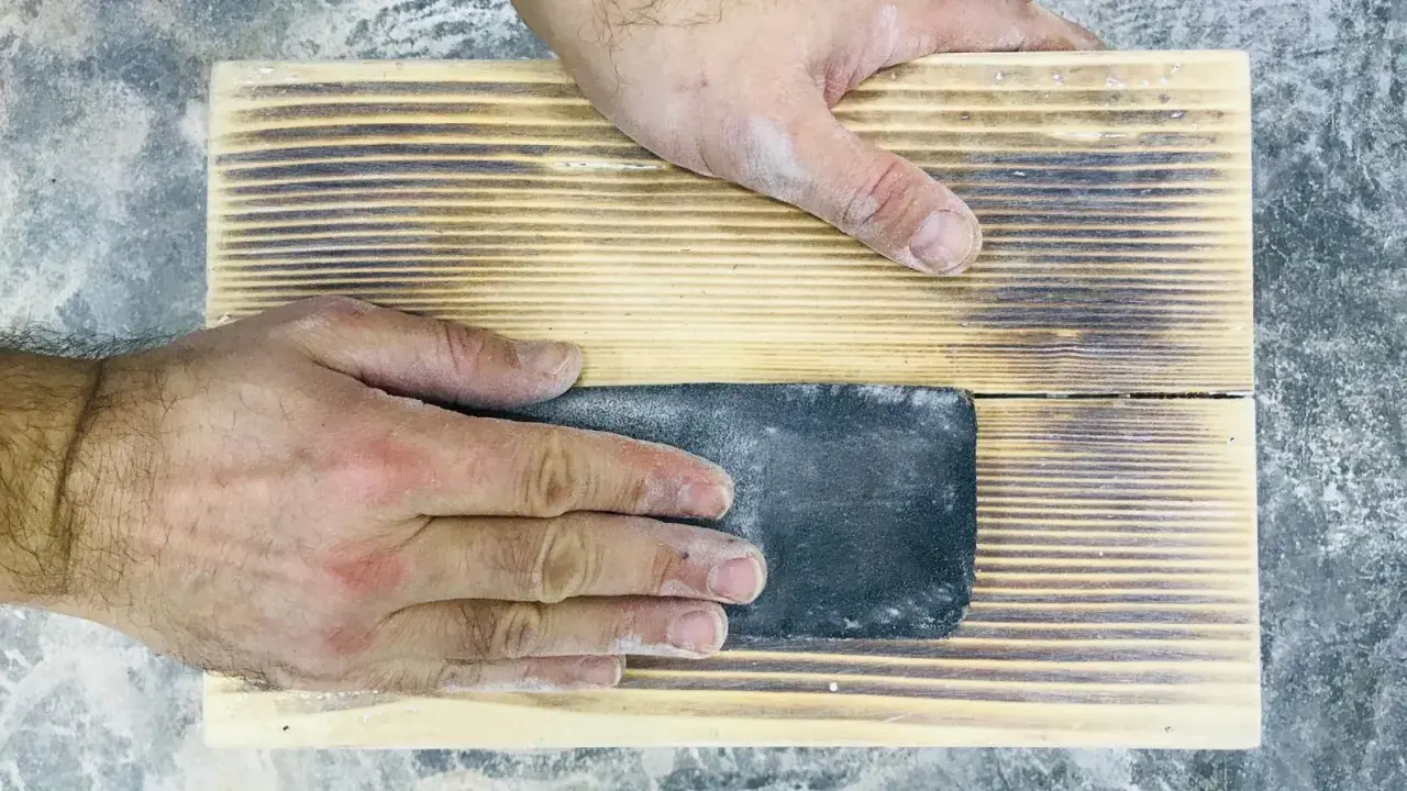 Mani che levigano un pezzo di legno con carta vetrata per sverniciare legno, rimuovendo la vernice vecchia.