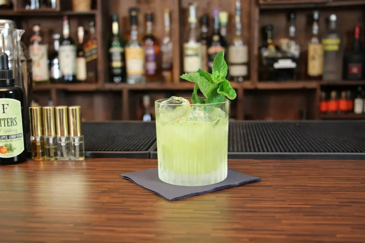 Ein grüner Tequila-Cocktail mit Minze und Eis auf einem Holztresen, im Hintergrund Regale mit Flaschen.