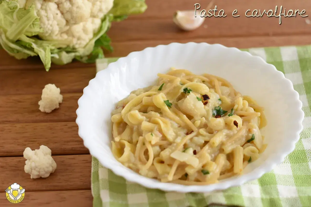 Un piatto di pasta e cavolfiore cremoso, guarnito con prezzemolo e peperoncino. Sullo sfondo, un cavolfiore fresco e uno spicchio d'aglio.