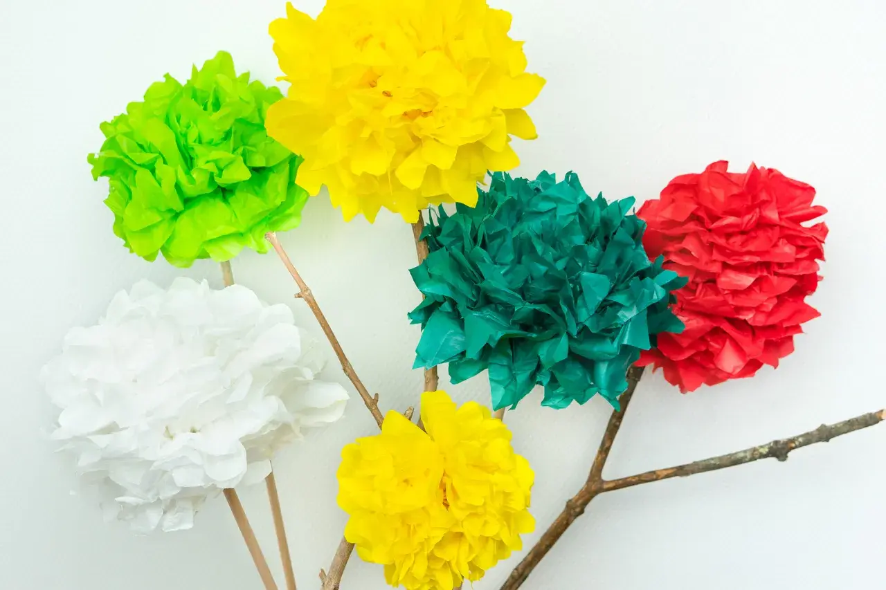 Bunte Seidenblumen basteln: Fünf Papierblumen in Rot, Grün, Gelb und Weiß auf Holzästen vor weißem Hintergrund.