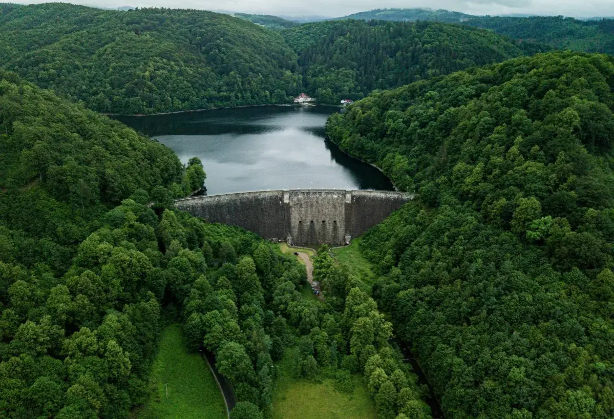 Zapora w górach Dolnego Śląska, otoczona gęstym, zielonym lasem. Woda w zbiorniku odbija pochmurne niebo.