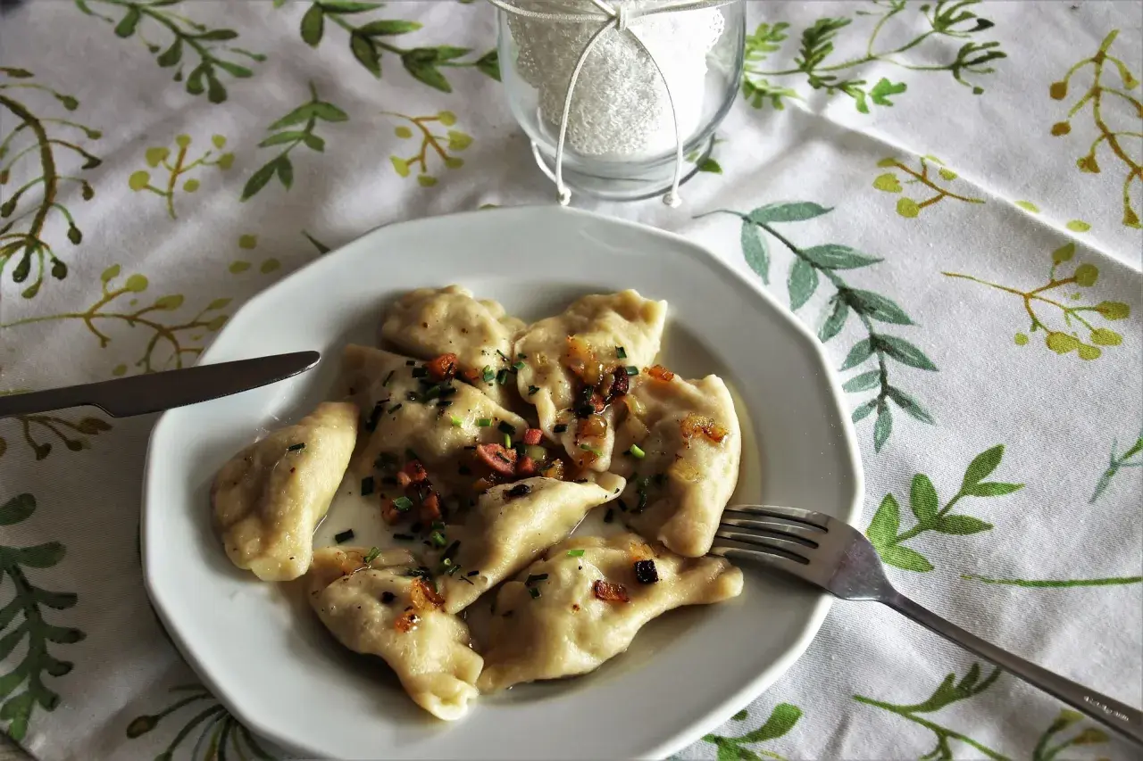 Pyszne pierogi ruskie z farszem Magdy Gessler, okraszone skwarkami i szczypiorkiem, na talerzu z widelcem i nożem.