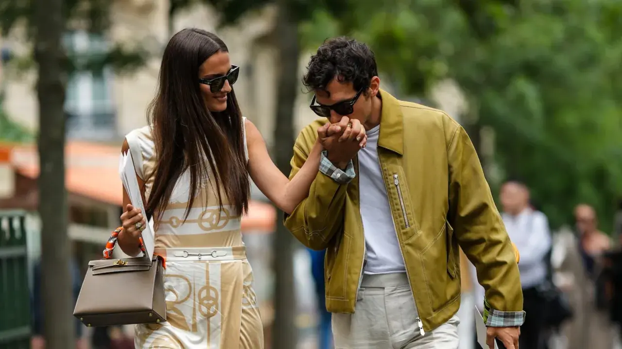 Ein Mann küsst die Hand einer Frau. Sein Outfit für ein erstes Date: gelbe Jacke, weißes T-Shirt, helle Hose. Sie trägt ein gemustertes Kleid und eine Tasche.
