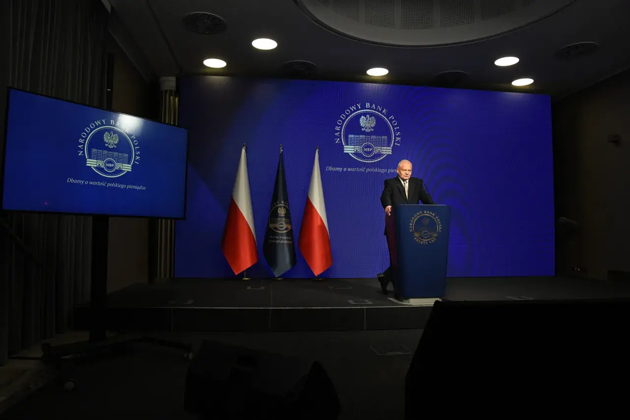 Prezes NBP Adam Glapiński przemawia na tle flag i ekranu z logo banku. Wiron 1m.