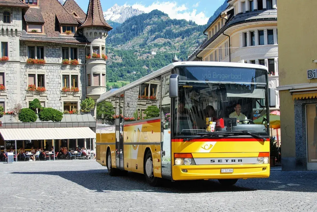 Żółty bus do Szwajcarii, z napisem "via Brigerbad", stoi na brukowanym placu przed kamiennymi budynkami i górami.