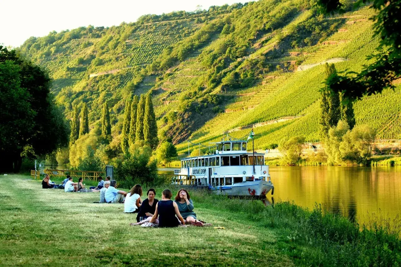 Entspannte Menschen genießen die Sonne am Ufer der Mosel. Ein Ausflugsschiff liegt vor, im Hintergrund steile Weinberge. Perfekte Mosel Ausflugsziele.