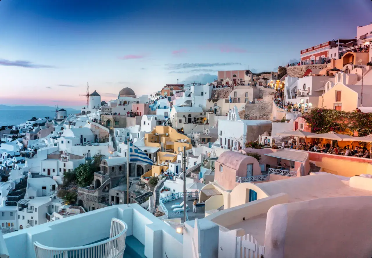 Santorini nocą, z białymi domkami, wiatrakami i grecką flagą, w blasku zachodzącego słońca.