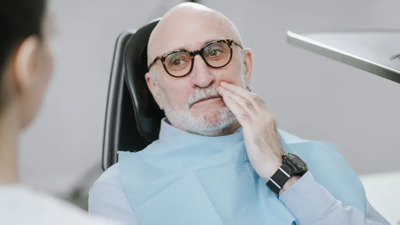 Starszy mężczyzna w okularach siedzi na fotelu dentystycznym, trzymając się za policzek. Wygląda na to, że boli go rozchwiany ząb.