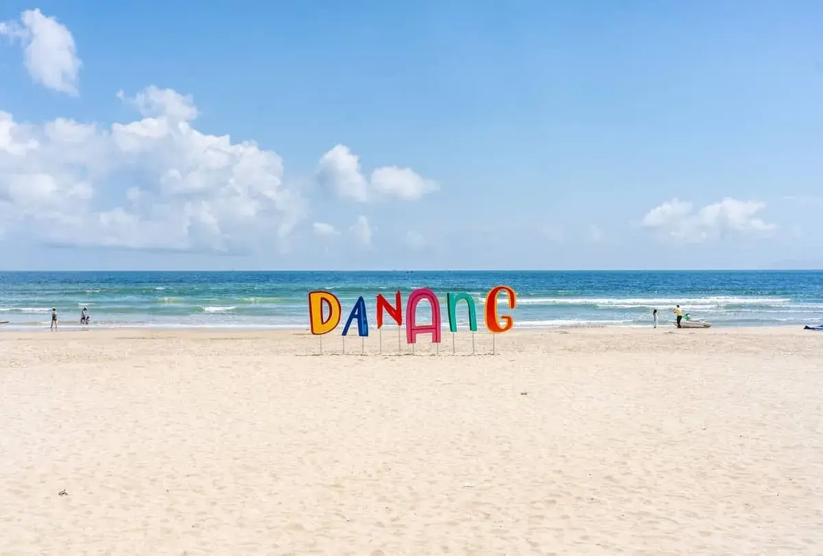 Kolorowe litery "DANANG" na piaszczystej plaży w Wietnamie, z błękitnym morzem i niebem w tle.