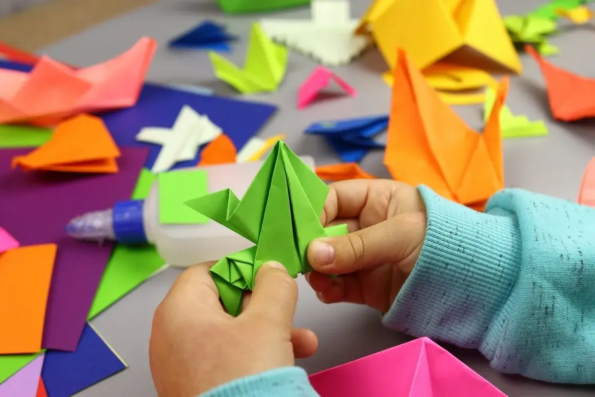 Dziecko tworzy proste origami z kartki a4. Wokół kolorowe figurki i klej.