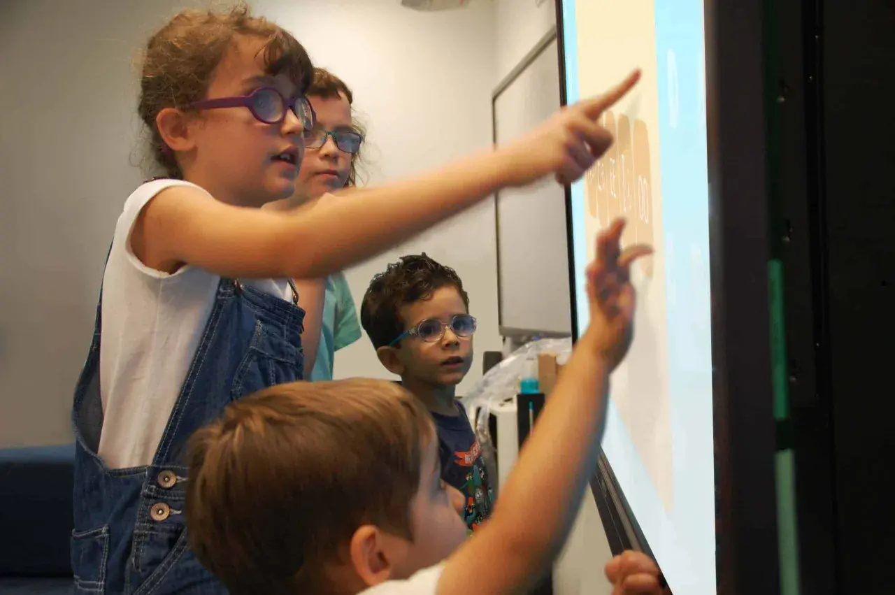 Kinder mit Brillen lernen interaktiv an einem Touchscreen. Sie zeigen auf Zahlen und Symbole, was multisensorisches Lernen ermöglicht.