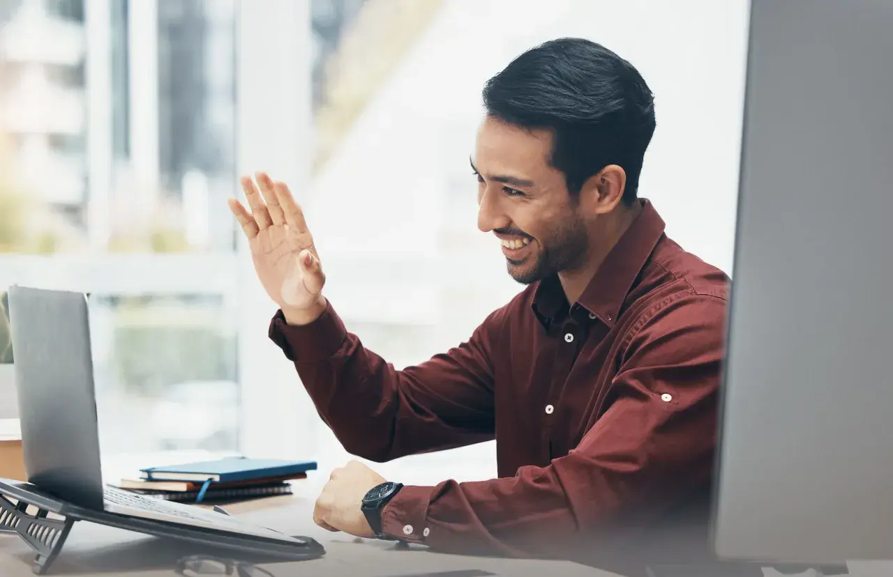Man waves hello during an online emotional intelligence coaching session, smiling warmly at his laptop.