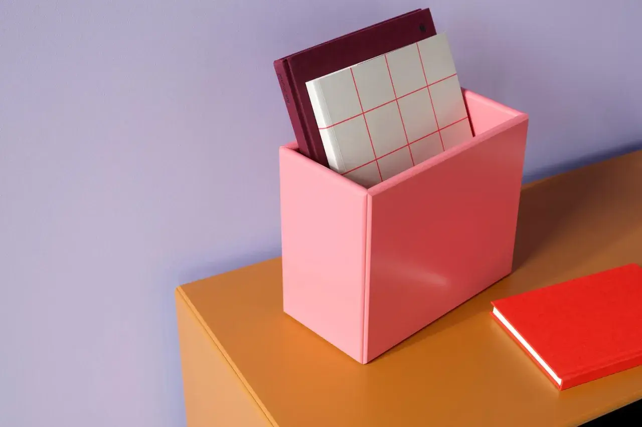 Ein rosa Behälter mit Büchern steht auf einem orangefarbenen Regal vor einer lila Wand. Welche Farbe passt zu lila? Hier sehen wir Rosa und Orange als passende Akzente.